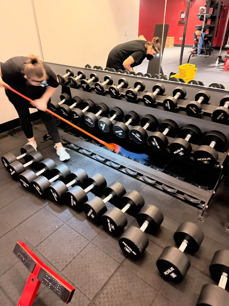 Two crew members cleaning the floor around dumbbell racks in a strength gym