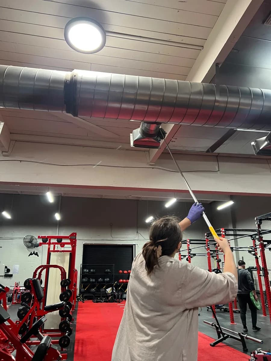 Crew member high dusting exposed ductwork during a gym deep clean in South Lake Union, Seattle