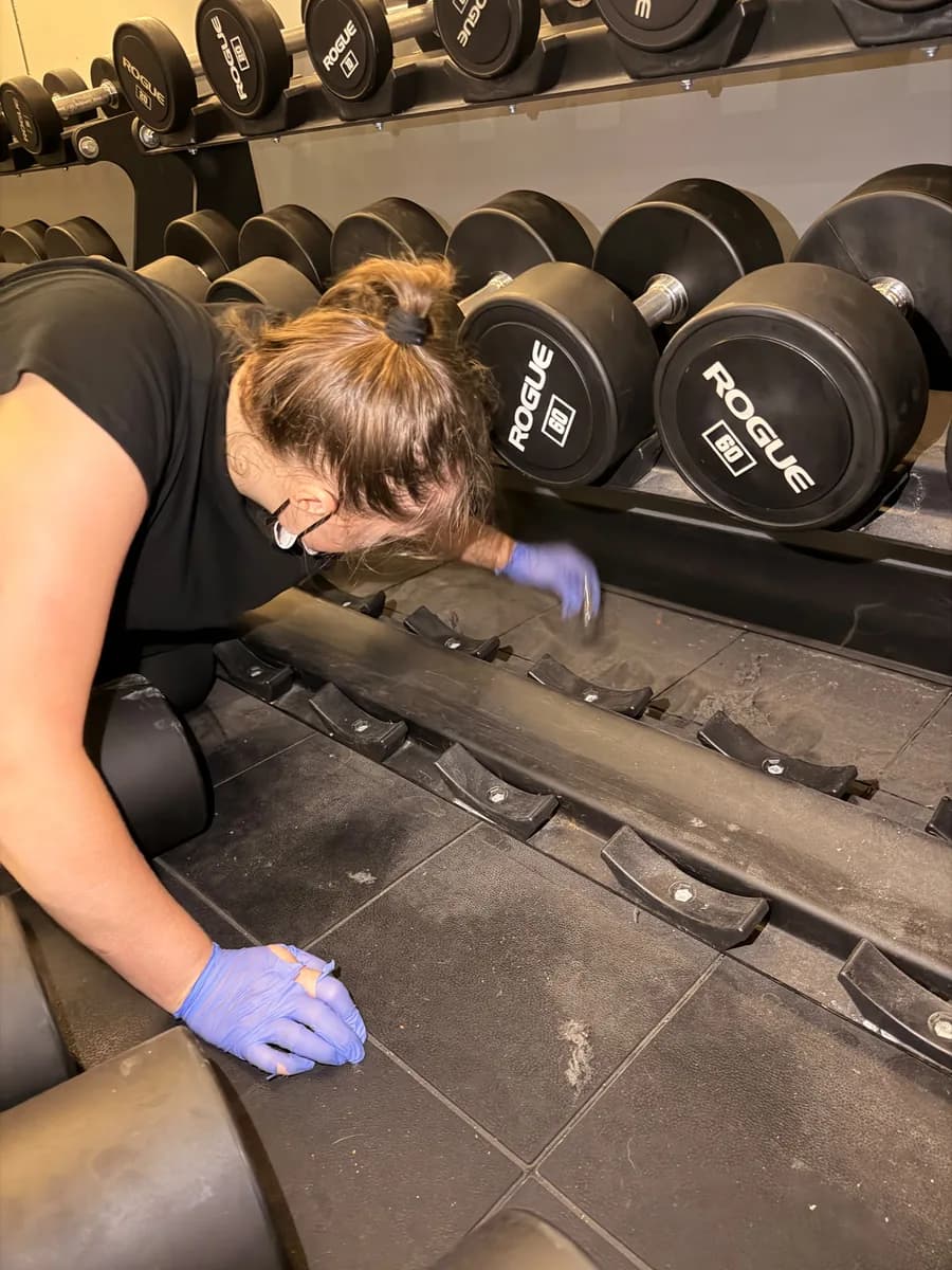 Crew member in gloves hand-cleaning the floor under a dumbbell rack